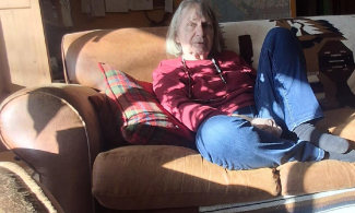 Heather Pritchard in a red sweater, seated on a brown couch with the afternoon sunlight streaming in.